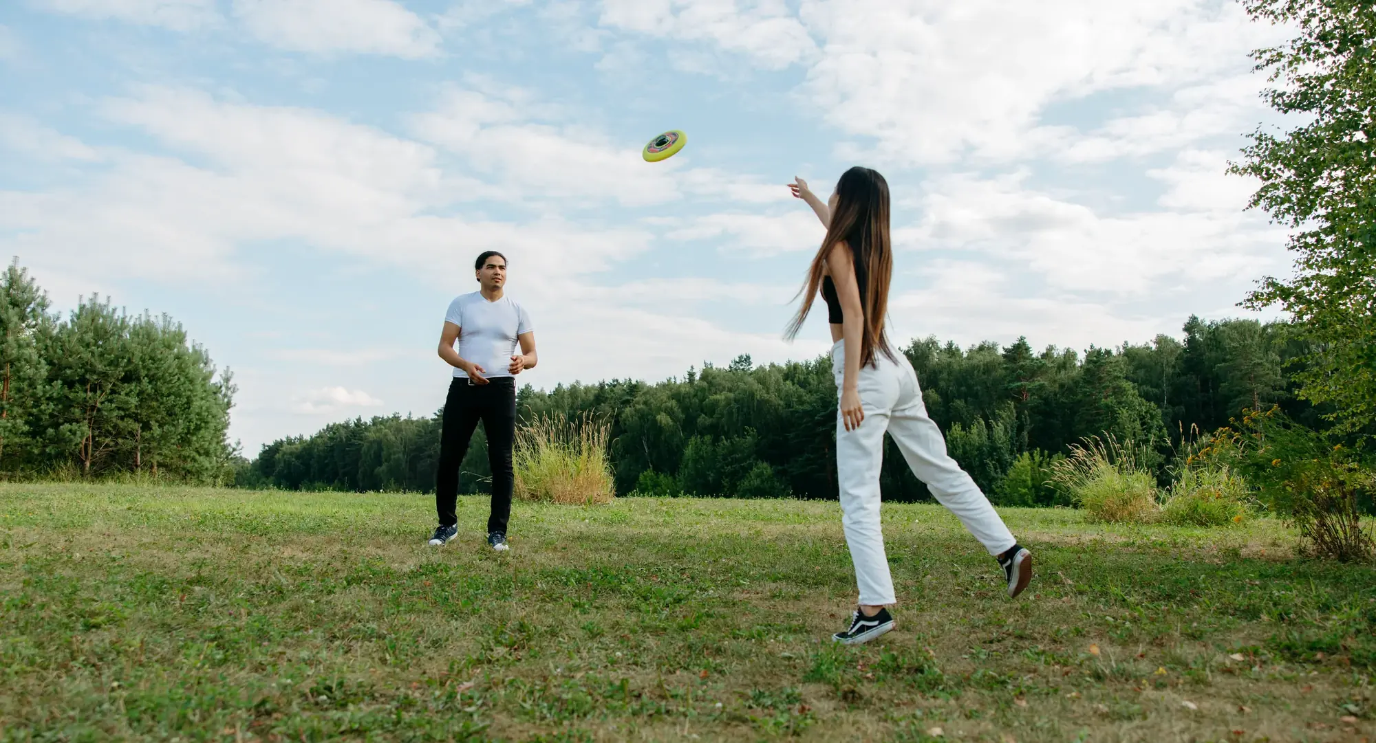 Two adults playing ultimate frisbee on an open grass field, disc mid-flight against a summer sky