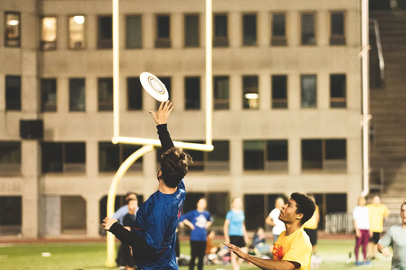 Ultimate frisbee players competing for a disc under floodlights at an evening match